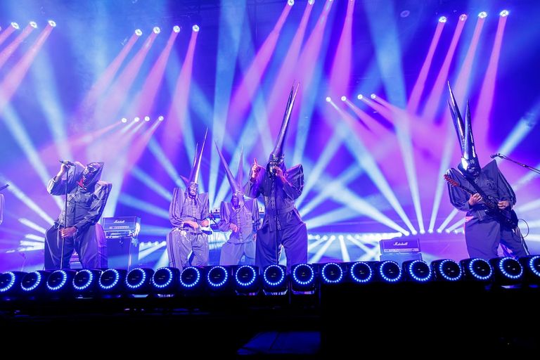 Australian rock band TISM performing live on stage in matching grey suits and pointed masks under dramatic purple lights.